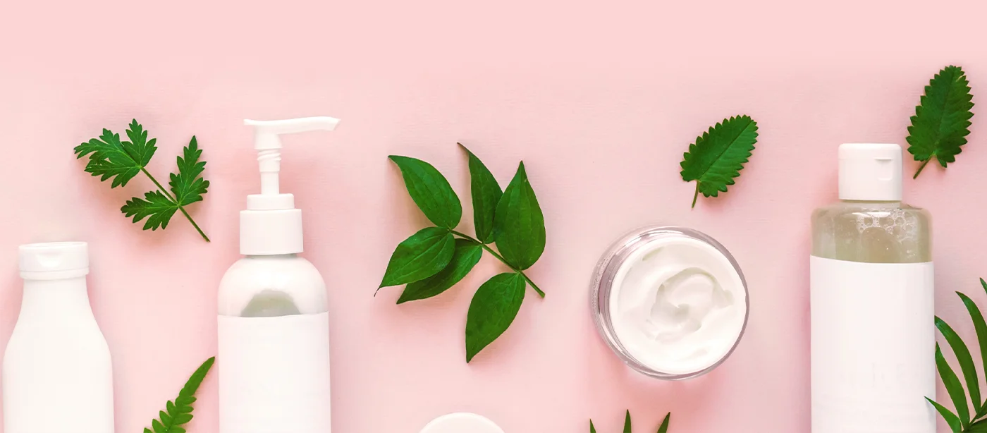 Assorted white containers of skincare products and green leaves arranged on a pink surface, emphasizing natural beauty theme.